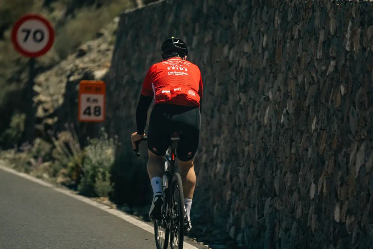 A cyclist in a red jersey climbs a sunny road through green hills.
